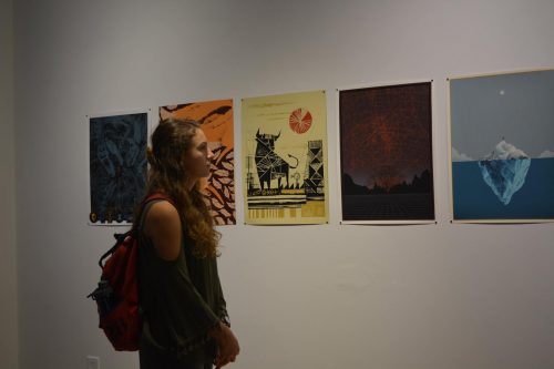 Woman with curly hair and backpack viewing colorful art prints displayed on white gallery wall