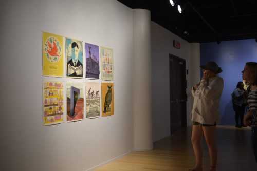 Visitors viewing colorful poster art exhibition on white gallery wall with track lighting and hardwood floors