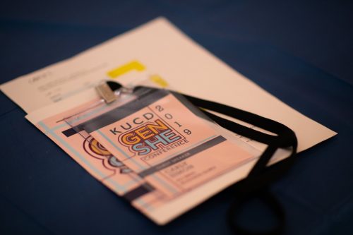 Smartphone displaying KUCD GenShed 2019 conference website on screen with paper documents underneath on dark surface