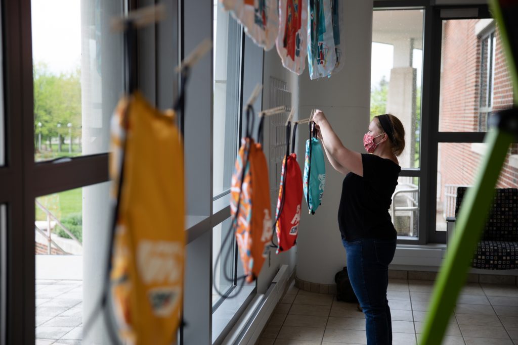 Woman wearing face mask hanging colorful laundry on indoor drying rack near large windows in modern home