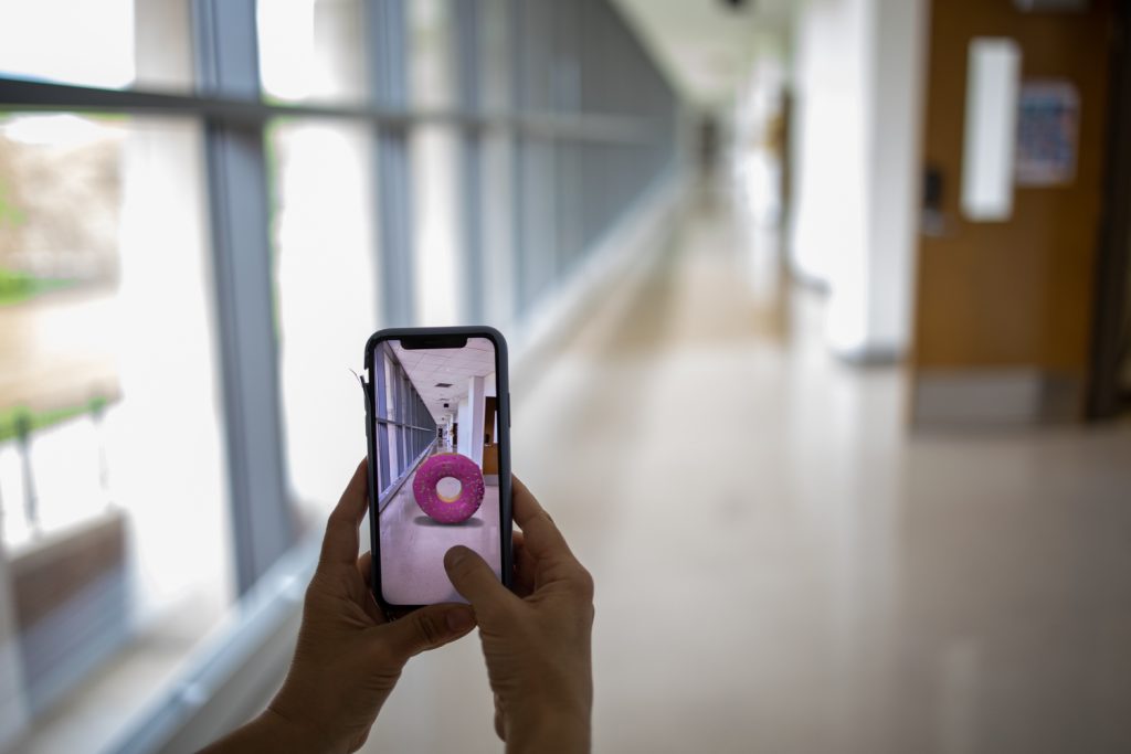 Person using augmented reality app on smartphone to view virtual pink donut in modern office hallway
