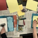 Two people doing printmaking art project with rollers, blue paint, yellow paper, and newspaper on work surface.
