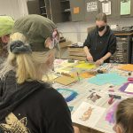 Students working on colorful art projects at table in classroom with art supplies and storage cabinets