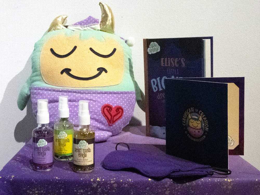 DreamDefenderz_Gallery Unicorn plush toy with spray bottles, sleep mask, and books on purple bedding - relaxation and self-care gift set