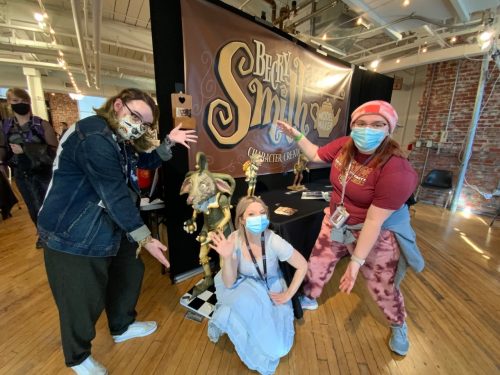 Three women in masks pose with fantasy creature puppets at Bear and Mythos character creation booth at convention