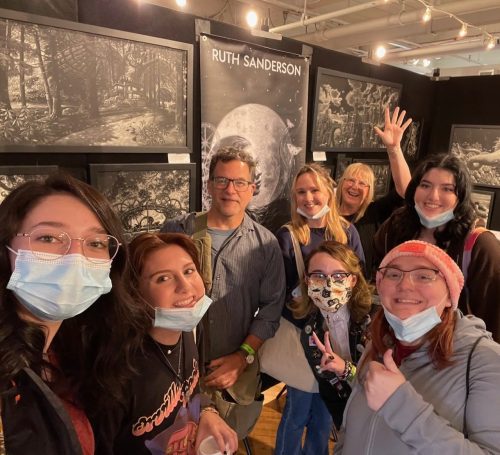 Group of people wearing masks posing together at Ruth Sanderson art exhibition with detailed fantasy artwork displayed