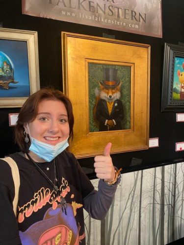 Woman with blue mask giving thumbs up next to framed fox portrait painting at Lisa Falkenstern art gallery exhibition