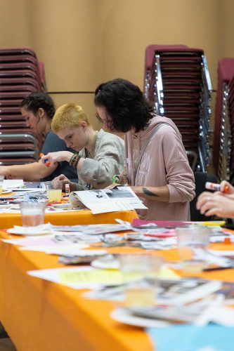 Students working on art projects at orange table with craft supplies and stacked chairs in background