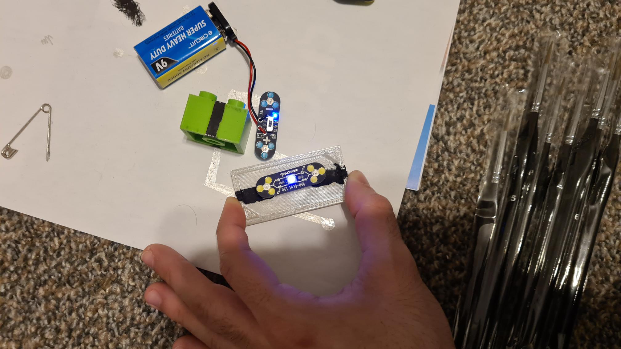 003-TestingLED Person holding 3D printed electronics project with circuit board, 9V battery, and LED components on white surface