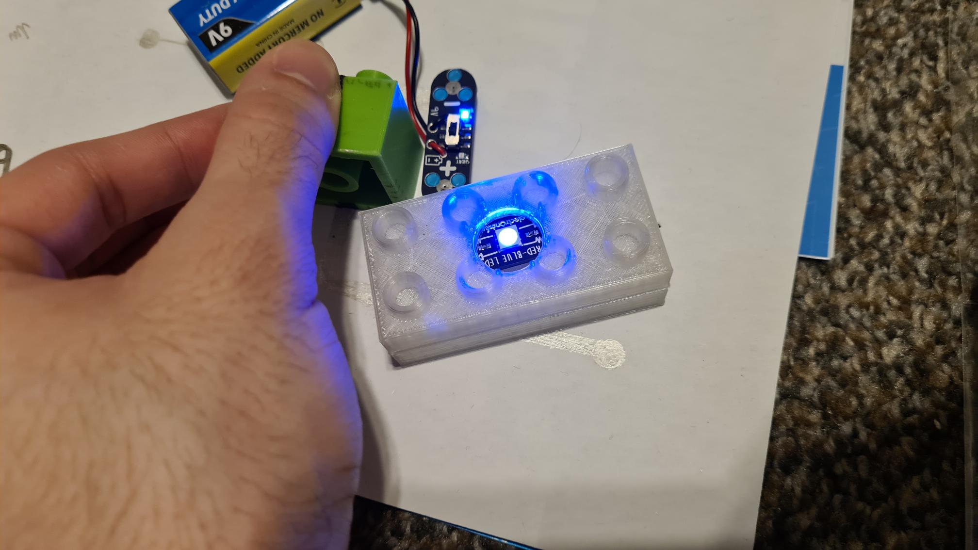 004-TestingFullCasing Hand holding UV light over 3D printed case with glowing blue circuit board and electronic components inside