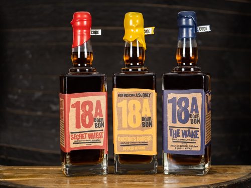 Three bottles of 18th Street Distillery bourbon whiskey with red, yellow and blue labels on wooden surface